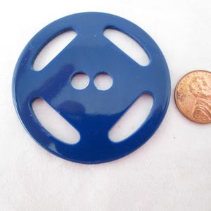May include: A round, blue button with a unique design, featuring four elongated openings and two smaller holes. A penny is placed next to the button for size comparison.