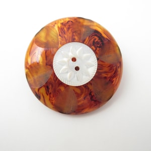 May include: A round button featuring a glossy, amber-colored tortoiseshell pattern on the outer ring and a circular white center inlay. The white center has a carved floral petal design and two central holes for attachment to a garment.