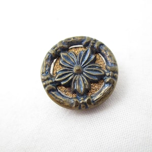 May include: A round, antique brass button with a floral design. The button features a central blue flower with detailed petals, surrounded by a textured gold background and a decorative outer ring. The button is set against a white fabric.