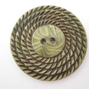 May include: A large, olive-green button with a spiral design. The button features a central, smaller circle with two holes for sewing. The outer edge has a textured, geometric pattern. The button is likely made of a durable material.
