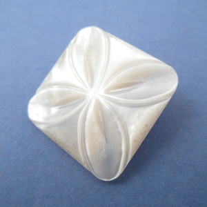 May include: A square-shaped button crafted from iridescent white mother-of-pearl, featuring a carved four-petal floral design on its surface. The button has a smooth, polished finish with natural pearlescent highlights and rounded corners.