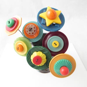 May include: A decorative arrangement of layered plastic buttons in various shapes and colors, including a yellow star, red, green, blue, orange, and yellow circular buttons, all mounted on thin dark wires to resemble a bouquet of flowers.