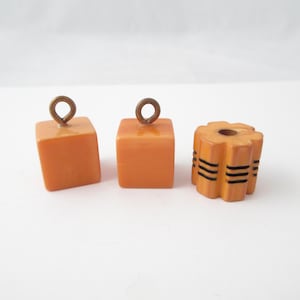 May include: Two orange cube-shaped beads with attached brass-toned metal loops and one orange cylindrical bead featuring a scalloped edge and horizontal black line details around the center of the piece.