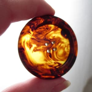 May include: A large, round button with a tortoiseshell pattern. The button is a mix of amber, brown, and yellow hues, with four small holes for sewing. The button is held between two fingers.