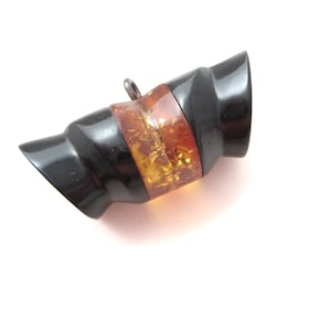 May include: A cylindrical pendant featuring two glossy black end sections and a central band of translucent amber-colored material with internal golden flecks. A small metal jump ring is attached to the top edge of the central section.