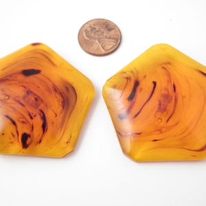 May include: Two pentagonal buttons featuring a translucent golden-yellow base with dark brown marbled swirls. A copper-colored United States penny is positioned between the buttons to provide a visual reference for their size and scale.