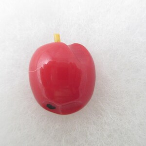 May include: A glossy red apple-shaped bead with a smooth, rounded surface. It features a small, short yellow stem protruding from the top and a single, distinct black dot located near the bottom edge of the red fruit-shaped object.