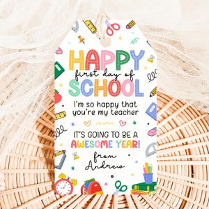 Puede incluir: Una colorida etiqueta de regalo con el texto "HAPPY first day of SCHOOL". La etiqueta incluye ilustraciones temáticas escolares como lápices, tijeras y libros. El mensaje dice "I'm so happy that you're my teacher. It's going to be a AWESOME YEAR!" y está firmado "from Andrew".