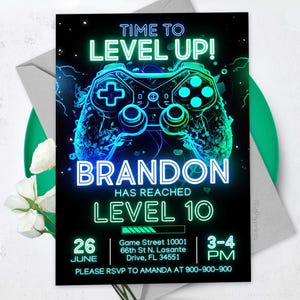 May include: A neon green and blue video game controller graphic on a black background with the text "Time to Level Up!" and "Brandon Has Reached Level 10". The invitation includes the date "June 26", time "3-4 PM", and location "Game Street 10001, 66th St N. Losante Drive, FL 34551".
