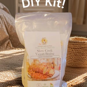 May include: A white plastic bag with a label that says "Slow Cook Vegan Braise" and "Plant Based Meal Kit". The bag is sitting on a woven rug. The label also has a picture of a vegan braise dish.