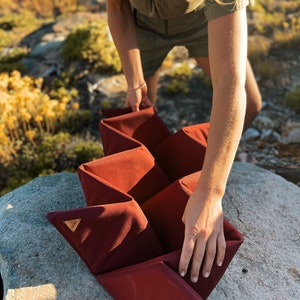 Pu&ograve; includere: Un contenitore portaoggetti bordeaux a fisarmonica con un design geometrico. Il contenitore &egrave; realizzato in un materiale morbido e strutturato e viene tenuto all'aperto. Una piccola etichetta triangolare in pelle &egrave; visibile.