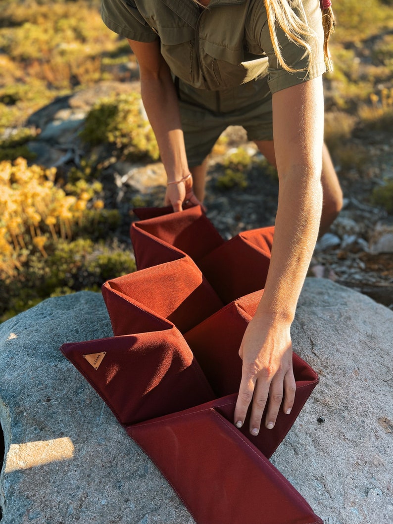 Pu&ograve; includere: Un contenitore portaoggetti bordeaux a fisarmonica con un design geometrico. Il contenitore &egrave; realizzato in un materiale morbido e strutturato e viene tenuto all'aperto. Una piccola etichetta triangolare in pelle &egrave; visibile.