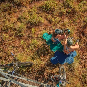 Pu&ograve; includere: Vista aerea di due persone con caschi e abbigliamento sportivo a fantasia, sedute su tappetini blu e verdi in un'area erbosa. Sono visibili mountain bike nelle vicinanze, suggerendo un tema di ciclismo o attivit&agrave; ricreative all'aperto.