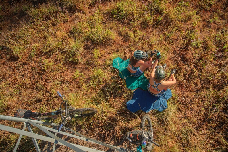 Pu&ograve; includere: Vista aerea di due persone con caschi e abbigliamento sportivo a fantasia, sedute su tappetini blu e verdi in un'area erbosa. Sono visibili mountain bike nelle vicinanze, suggerendo un tema di ciclismo o attivit&agrave; ricreative all'aperto.