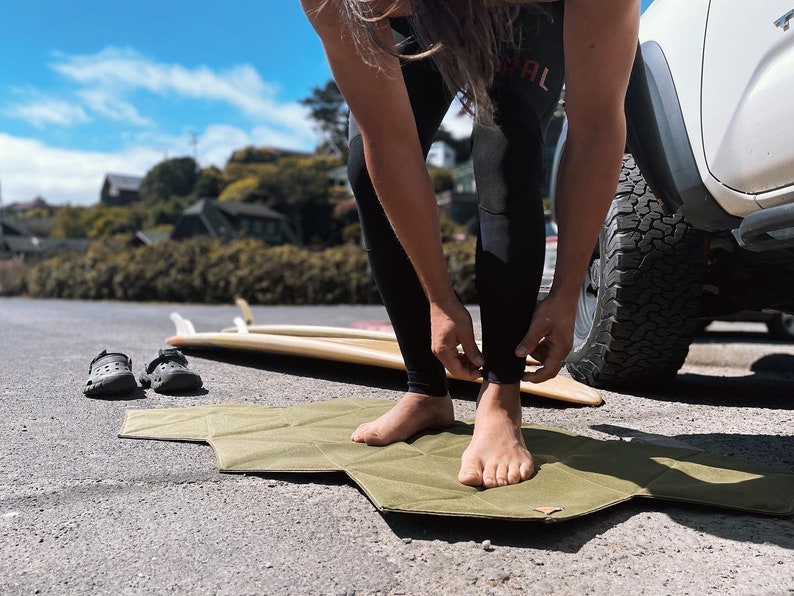 Pu&ograve; includere: Un tappetino verde oliva &egrave; appoggiato sul marciapiede. Una persona si sta sistemando i pantaloni della muta neri. Due tavole da surf e un paio di zoccoli neri sono sullo sfondo. La scena &egrave; all'aperto in una giornata di sole.