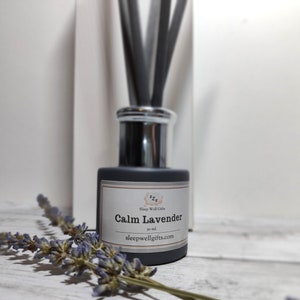 May include: A grey glass diffuser bottle with black reeds and a white label that reads "Calm Lavender" with the text "Sleep Well Gifts" and the website address "sleepwellgifts.com". The bottle is sitting on a white wooden surface with dried lavender sprigs.