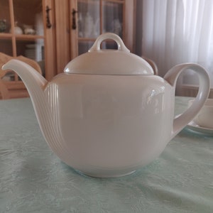 Pode incluir: Uma chaleira de porcelana branca com tampa e um bico curvo. A chaleira está sobre uma toalha de mesa azul claro.