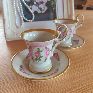Puede incluir: Dos tazas de espresso de porcelana blanca con platos a juego, con un patrón de rosas rosas, un motivo de lazo rosa y bordes dorados en los bordes y las asas. Cada taza tiene un asa curva y se asienta sobre un plato decorado con rosas.