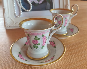 May include: Two white porcelain espresso cups and matching saucers featuring a pink rose floral pattern, a pink ribbon motif, and gold trim on the rims and handles. Each cup has a curved handle and sits on a saucer decorated with pink roses.