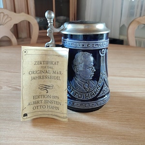 May include: A blue and white ceramic beer stein with a portrait of Otto Hahn and the text "Otto Hahn 1879" and "E=mc2". The stein is accompanied by a certificate for the "Original M&R. Jahressiedel Edition 1979 Albert Einstein - Otto Hahn".