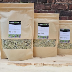 May include: Three brown paper bags of dried senna leaf, each with a clear window. The bags are labelled with "HERBSHOPUK" and "SENNA LEAF". The bags are on a wooden surface, with a brick wall in the background. The bags vary in size.