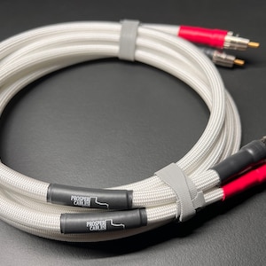 Puede incluir: Cables de audio trenzados blancos con conectores rojos y negros. Los cables están enrollados y asegurados con correas grises. Los extremos tienen conectores plateados y dorados. El texto "PROSPER CABLES" está impreso en los extremos negros de los cables.
