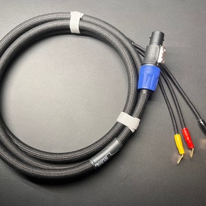 Puede incluir: Cable de audio negro con una textura tejida, asegurado con cinta blanca. Cuenta con un conector negro y azul y tres cables con puntas amarillas, rojas y negras. El cable está etiquetado con la palabra "PROSPER".