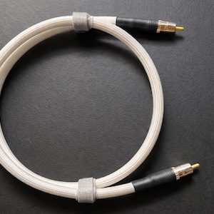 May include: A coiled audio cable featuring a white braided exterior, black heat-shrink tubing at the ends, and gold-plated RCA connectors. The cable is secured with two small grey fabric straps to maintain its circular shape for storage.