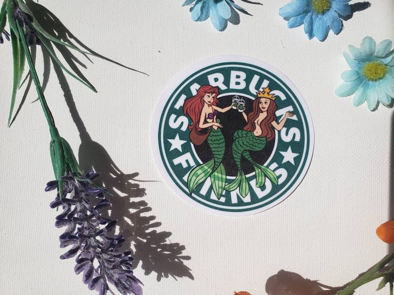 May include: Green and white sticker with the Starbucks logo and the text "Starbucks Friends" in a circle. Two mermaids are holding cups of coffee. One mermaid has red hair and the other has brown hair.
