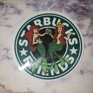 May include: A green and white sticker with the Starbucks logo and the word "Friends" written around it. Two mermaids are holding cups of coffee, one with red hair and one with blonde hair.