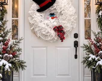 May include: A white mesh snowman wreath featuring a black top hat with a red band and green holly leaves, a painted snowman face with a carrot nose, and a red and black plaid ribbon bow attached to the bottom of the circular wreath.