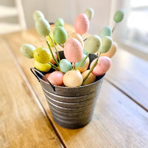 May include: A galvanized metal bucket filled with pastel-colored Easter egg decorations. The eggs are on stems and include shades of pink, green, yellow, and blue. The bucket sits on a wooden table, creating a rustic and festive display.