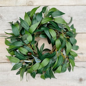 May include: A green olive branch wreath with small green olives. The wreath is made of artificial leaves and is perfect for decorating a door or wall.