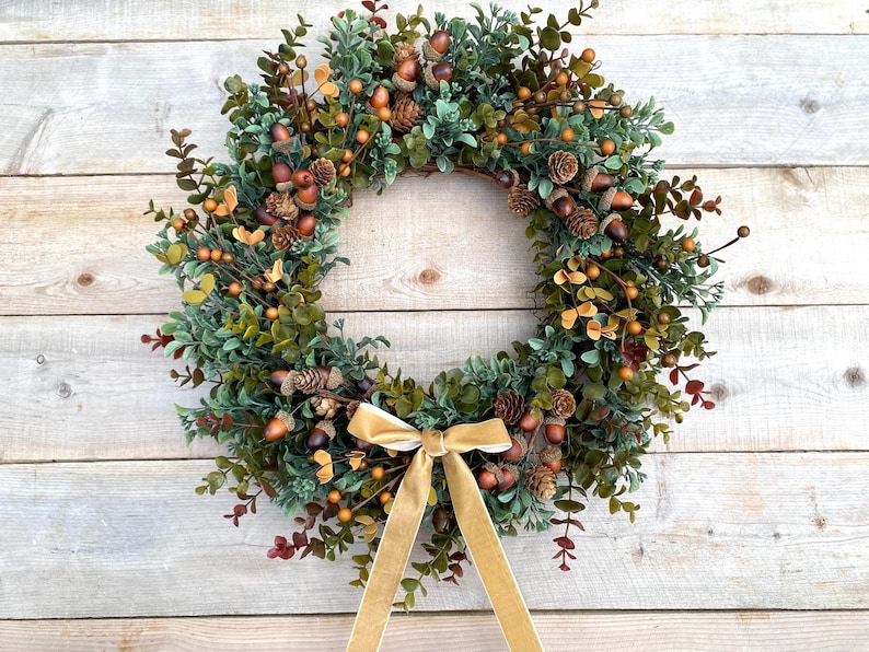 May include: A green, brown, and gold wreath with a gold velvet ribbon. The wreath is decorated with faux greenery, acorns, and pine cones.