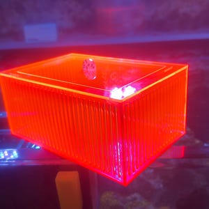 Pu&ograve; includere: Una scatola rettangolare arancione brillante con un esterno a coste. La scatola &egrave; illuminata da una luce arancione brillante, creando un display vibrante e accattivante. La scatola &egrave; probabilmente realizzata in un materiale traslucido.