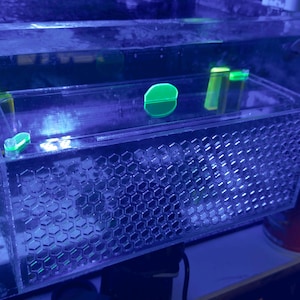 Pu&ograve; includere: Un filtro per acquario rettangolare trasparente con un motivo a nido d'ape sulla parte anteriore. All'interno del filtro ci sono diversi piccoli oggetti dai colori vivaci. L'acquario &egrave; pieno d'acqua e illuminato con luci blu e verdi.