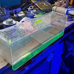 Pu&ograve; includere: Un contenitore rettangolare di plastica trasparente con un motivo a rombi sui lati e una base verde. Il contenitore &egrave; parzialmente riempito con un liquido trasparente. Il contenitore &egrave; su un banco di lavoro disordinato.
