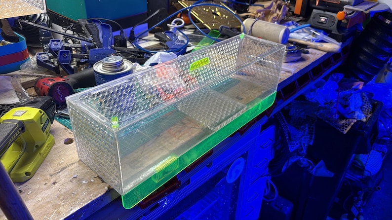 Pu&ograve; includere: Un contenitore rettangolare di plastica trasparente con un motivo a rombi sui lati e una base verde. Il contenitore &egrave; parzialmente riempito con un liquido trasparente. Il contenitore &egrave; su un banco di lavoro disordinato.