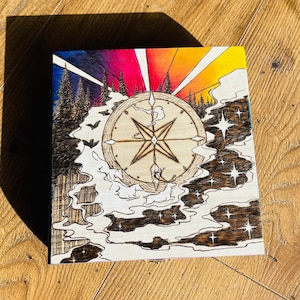 May include: A wooden box with a woodburned design of a compass, stars, and clouds. The compass is surrounded by a rainbow-colored sky and a forest.