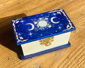 Puede incluir: Una pequeña caja de madera con una tapa azul decorada con estrellas doradas, lunas y flores blancas. La caja tiene un cierre dorado.