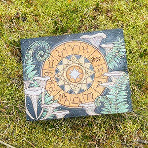 May include: A wooden plaque with a black background and a gold zodiac wheel with the signs of the zodiac. The plaque is surrounded by white mushrooms and green ferns.