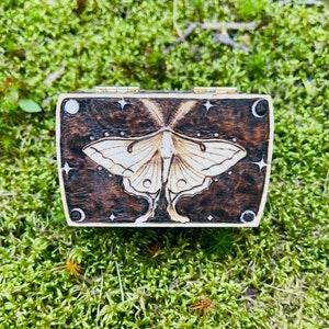 May include: A small wooden box with a hinged lid. The box is decorated with a woodburned design of a luna moth with a crescent moon and stars. The box is closed and sitting on a bed of green moss.