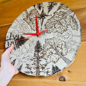 May include: A wood slice clock with a red hour hand and black minute and second hands. The clock face features a woodburned design of trees and a compass rose.