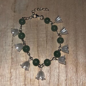 May include: A silver-toned chain bracelet featuring alternating dark green spherical beads and translucent white bell-shaped flower charms. The bracelet includes a metal lobster claw clasp and an adjustable extender chain with circular links.