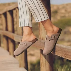 Könnte beinhalten: Taupefarbene Wildleder-Clogs mit Korkfußbett und Schnallen-Detail. Das offene Design hat eine runde Spitze und eine braune Korksohle. Diese lässigen Clogs sind perfekt für den Alltag.