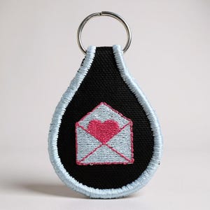 May include: Black teardrop-shaped keychain with a light blue embroidered border. The keychain features a light blue envelope design with a pink heart in the center. A silver key ring is attached at the top.