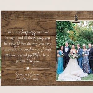 May include: A rustic wooden sign with a photo of a wedding party in a garden setting. The sign has a white inscription that reads "For all the happiness you have brought and all the lessons you have taught. For the way you have cared and the wisdom you shared. We are beyond grateful to have parents like you." The sign is signed "Simon and Dianna" and dated "October 30, 2022".
