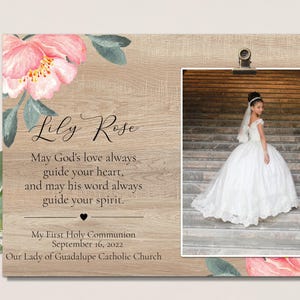 May include: A light wood photo frame with a white background and a floral design. The frame has a clip at the top to hold a photo. The photo shows a young girl in a white dress standing on a set of stairs. The text "Lily Rose" is written in black cursive font. The text "May God's love always guide your heart, and may his word always guide your spirit." is written in black font. The text "My First Holy Communion September 16, 2022 Our Lady of Guadalupe Catholic Church" is written in black font.