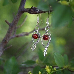 Peut inclure: Paire de boucles d'oreilles artisanales en argent, chacune ornée d'une perle rouge. Les boucles sont suspendues à des crochets argentés et se détachent sur un fond vert flou de feuilles et de branches.
