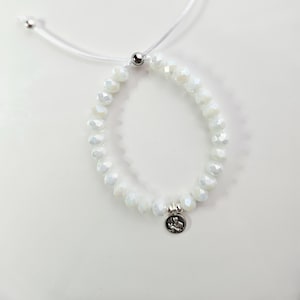 May include: White beaded bracelet with a small silver charm. The bracelet features iridescent, faceted beads strung on a white cord. The charm has a detailed design. The bracelet is adjustable.
